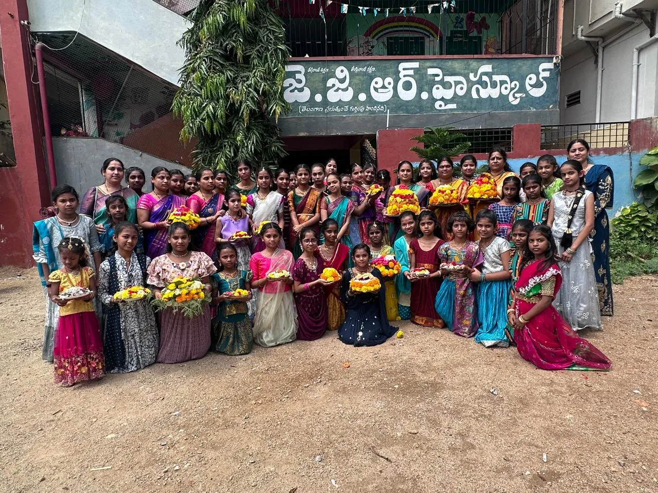Girls and teachers holding Bathukamma flower towers in front of the school